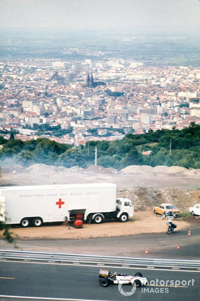 Jackie Oliver, BRM P153, pasa junto a un camión del Servicio Médico del Gran Premio Internacional, con una vista de la ciudad de Clermont-Ferrand al fondo.